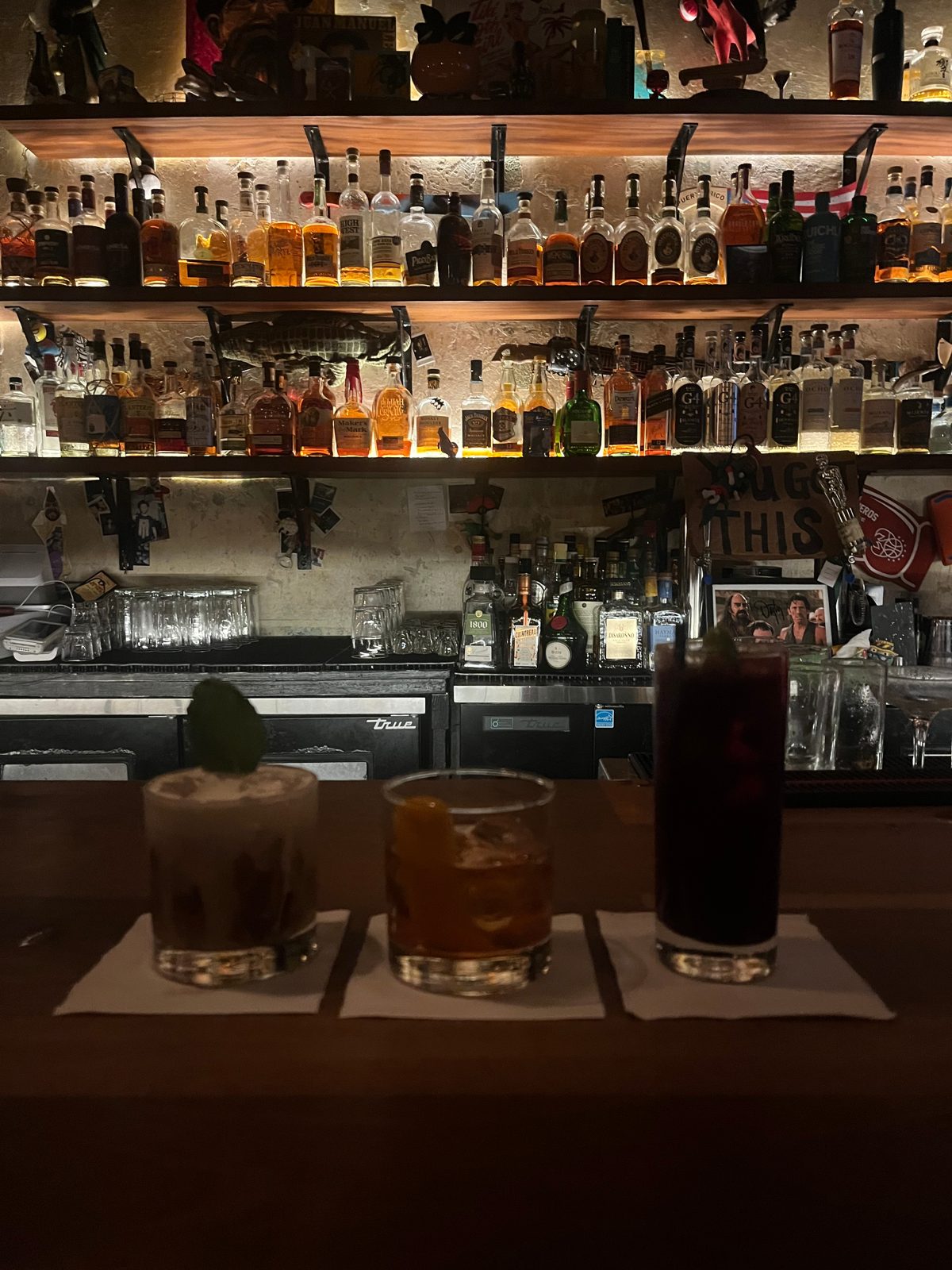 Three cocktails lined up on a bar, with shelves of bottles behind
