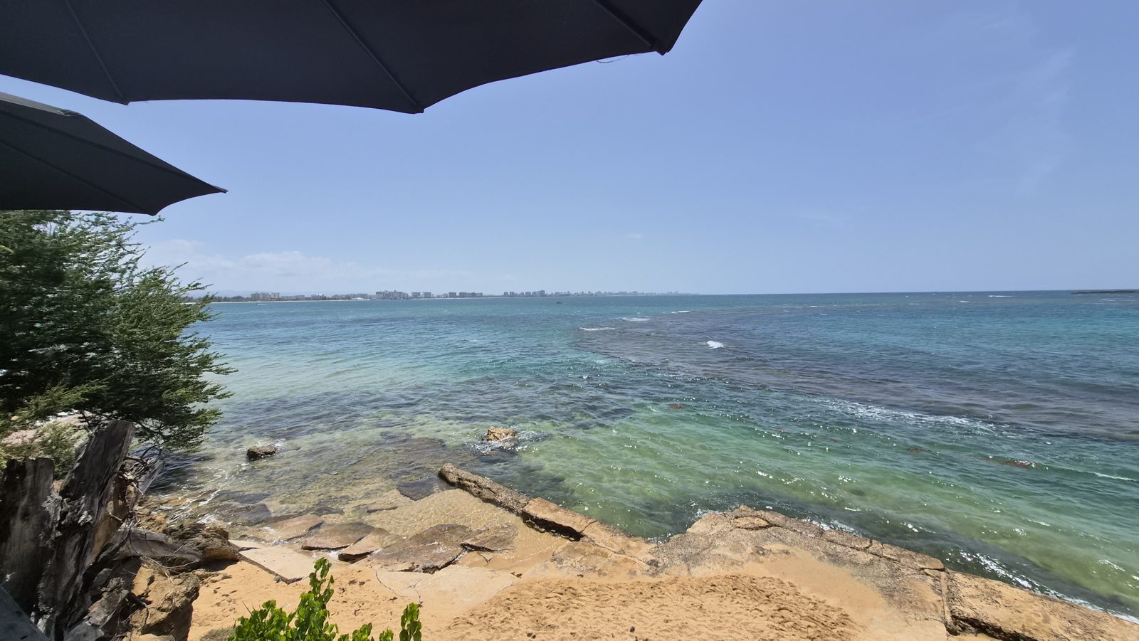 Clear turquoise water over flat coastal rock, seen from beneath a shaded terrace, with the city far across the bay