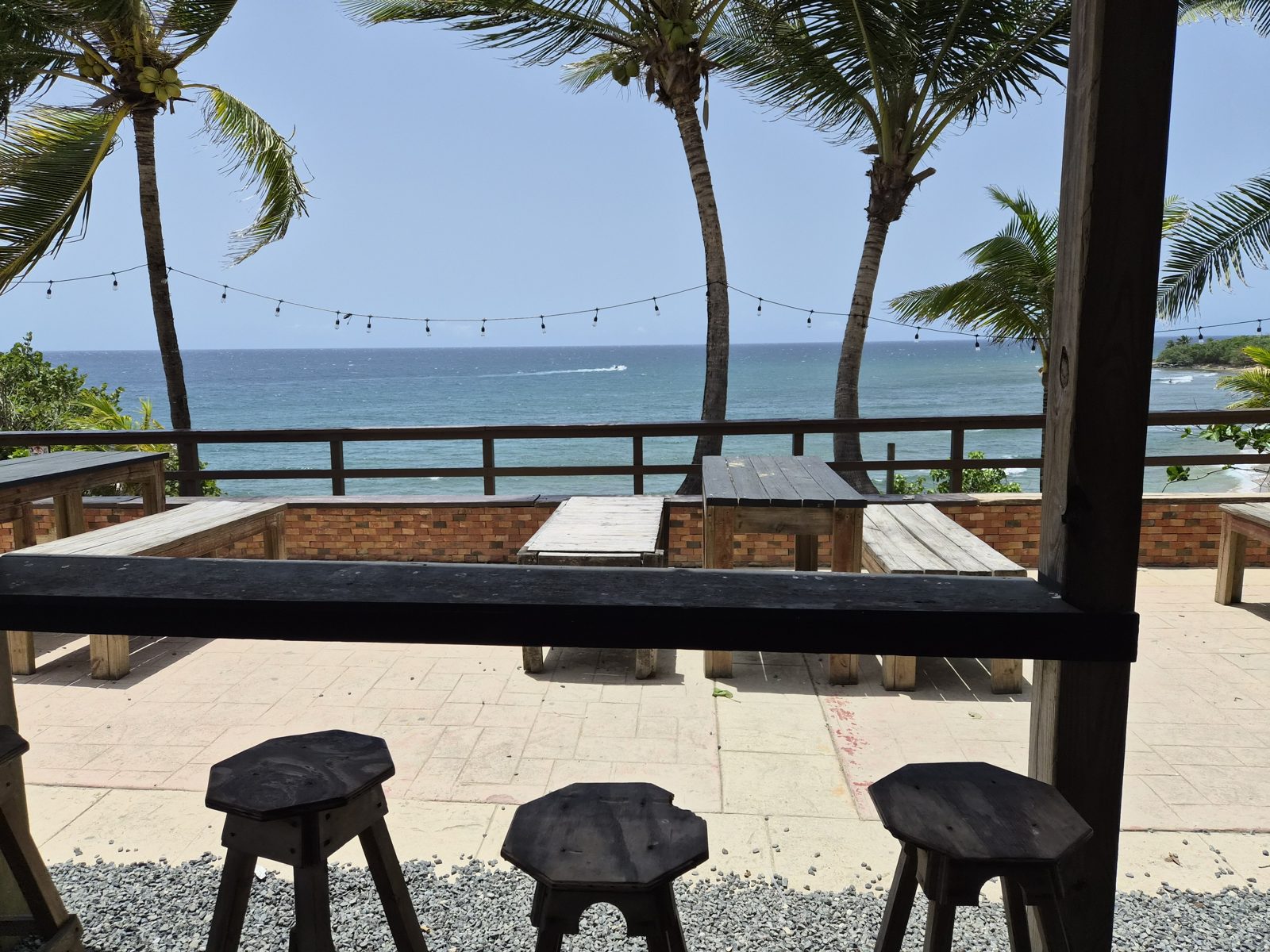 A shaded wooden terrace with stools and tables, looking out between palm trees to the sea